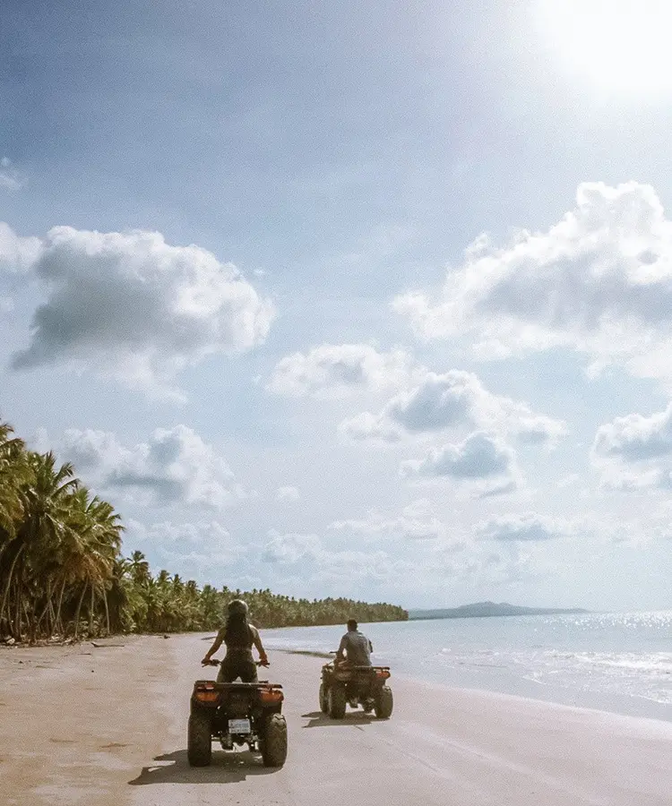 Two people riding atvs on a beach near the ocean.