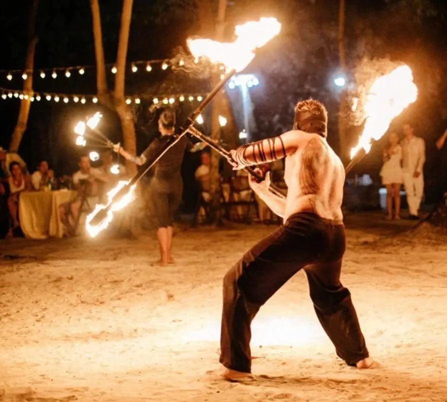 A man holding two fire staffs while standing on sand.