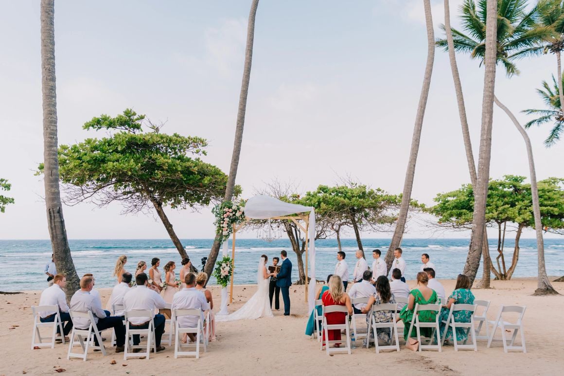 A beach wedding with many people sitting on the sand.